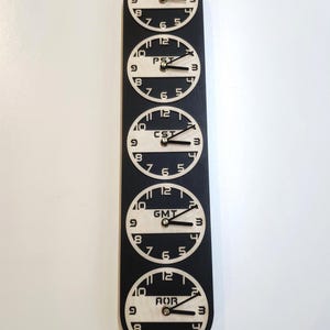 May include: A black and white wall clock displaying five different time zones: EST, PST, CST, GMT, and AOR. Each clock face is circular with black hands and numerals against a light background. The clocks are arranged vertically.
