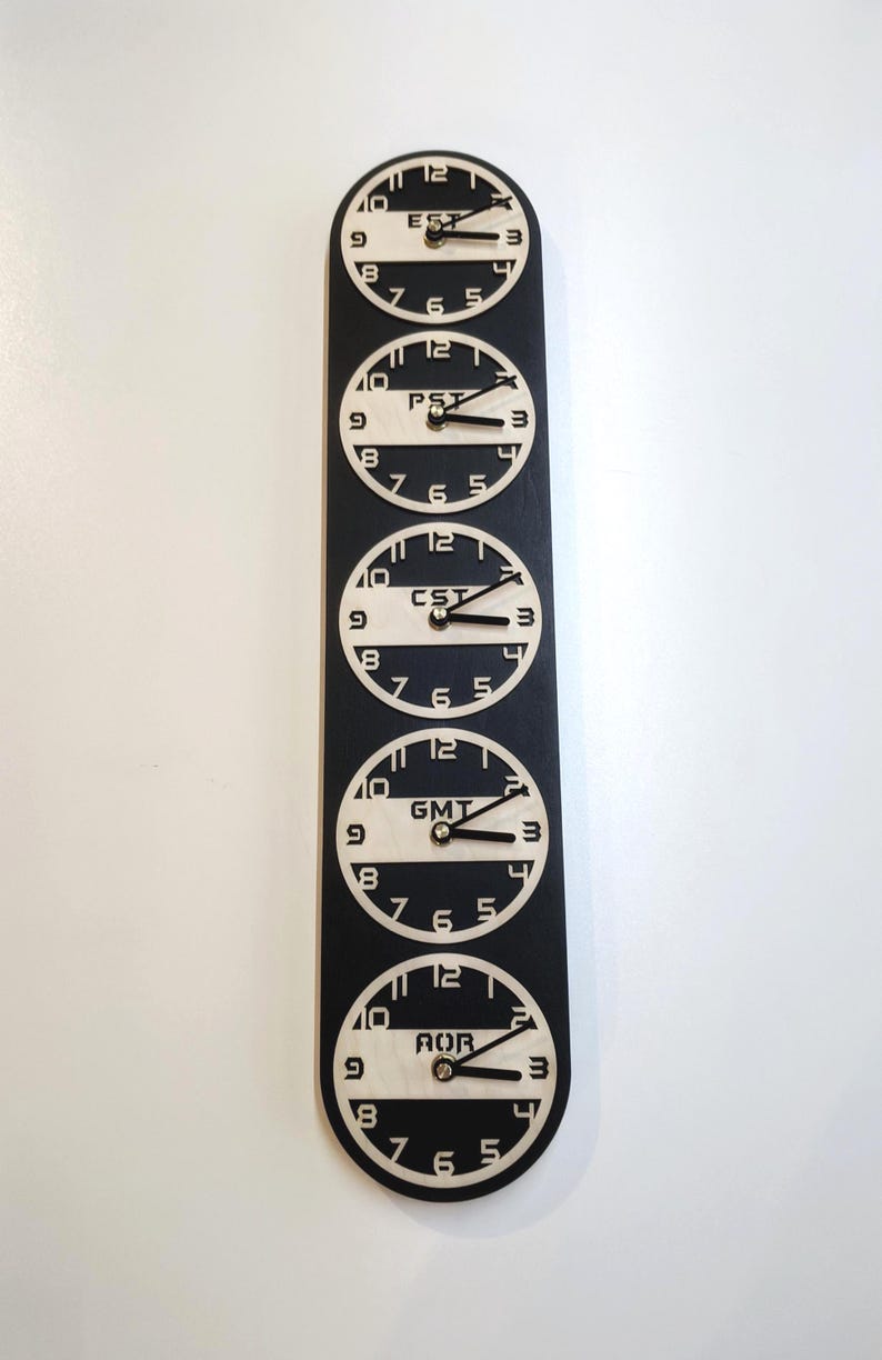 May include: A black and white wall clock displaying five different time zones: EST, PST, CST, GMT, and AOR. Each clock face is circular with black hands and numerals against a light background. The clocks are arranged vertically.