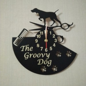 May include: Black metal wall clock with a silhouette of a dog, scissors, and a comb. The clock face has gold numbers and hands. The clock is shaped like a half circle and has the text "The Groovy Dog" on it.