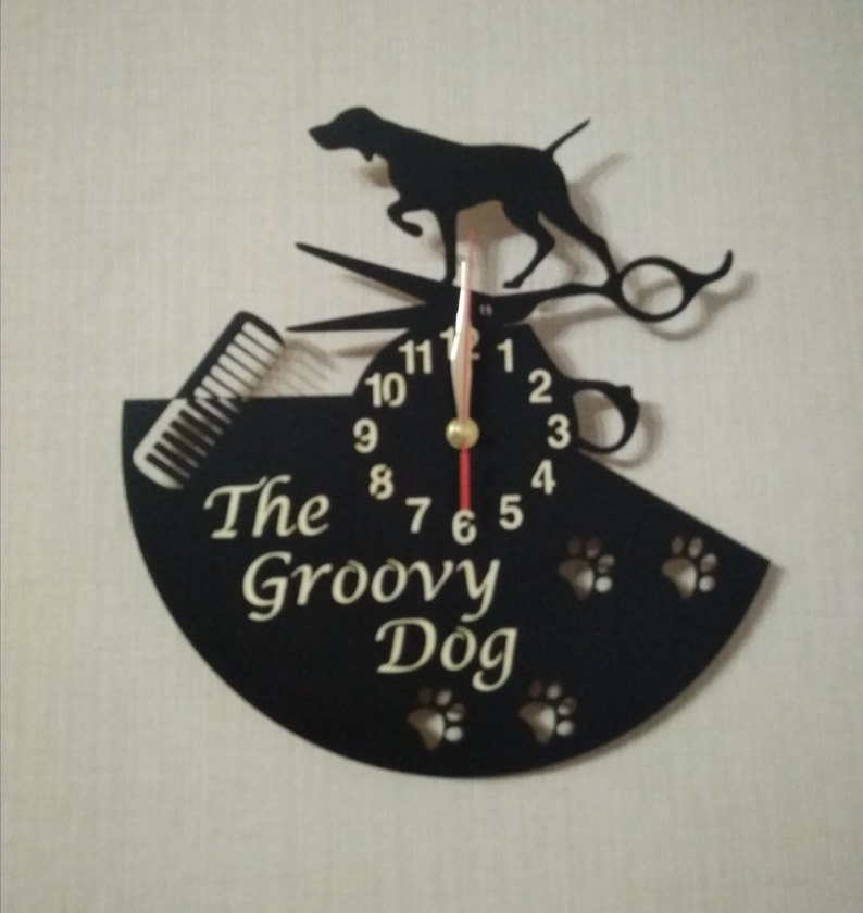 May include: Black metal wall clock with a silhouette of a dog, scissors, and a comb. The clock face has gold numbers and hands. The clock is shaped like a half circle and has the text "The Groovy Dog" on it.