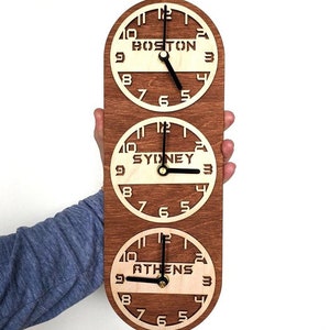 May include: A wooden wall clock with three circular faces, each displaying a different time zone: Boston, Sydney, and Athens. The clock has a dark brown frame and light wood clock faces with black hands. The clock is held against a white background.