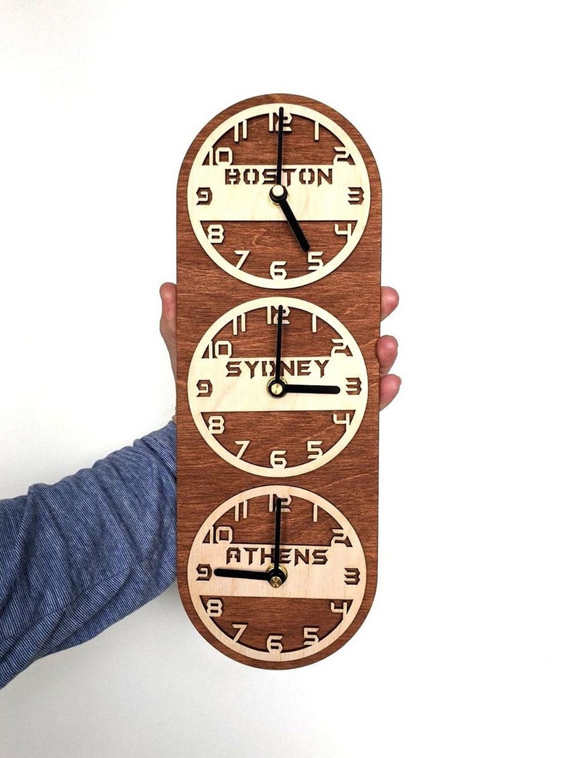 May include: A wooden wall clock with three circular faces, each displaying a different time zone: Boston, Sydney, and Athens. The clock has a dark brown frame and light wood clock faces with black hands. The clock is held against a white background.