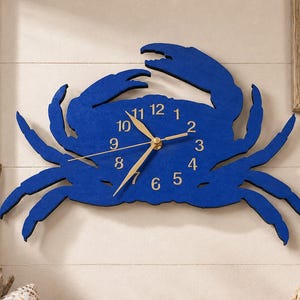 May include: A wall clock shaped like a crab with a deep blue finish. The clock face is integrated into the crab's body and features gold-colored numbers from 1 to 12, along with matching gold-colored hour, minute, and second hands at the center.