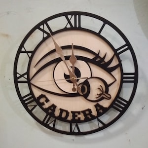 May include: A black and white clock with a large eye design and the word "CADERA" written in a decorative font. The clock has Roman numeral markings.