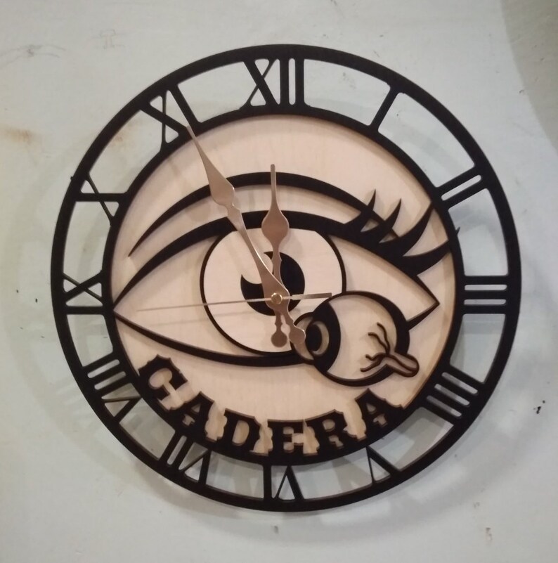 May include: A black and white clock with a large eye design and the word "CADERA" written in a decorative font. The clock has Roman numeral markings.