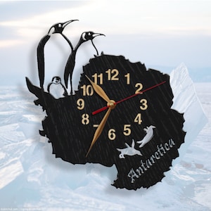 May include: A black wooden clock shaped like the continent of Antarctica with a gold clock face and black hands. The clock is surrounded by three penguins, two of which are standing on the top of the clock and one is sitting on the side. The clock face has the numbers 1 through 12 and the word "Antarctica" is written below the clock face.
