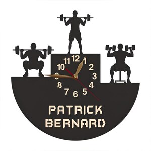 May include: Black and white silhouette of a clock with a weightlifting theme. The clock face has black numbers and a gold hand. The clock is surrounded by three weightlifters. The clock face has the name "PATRICK BERNARD" printed below it.