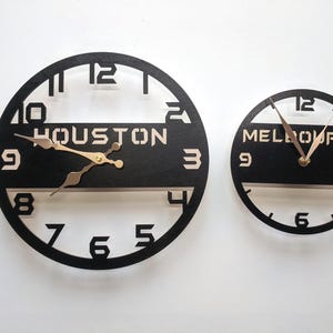 May include: Two black, circular wall clocks. One clock reads "HOUSTON" and the other reads "MELBOURNE" in white lettering. The clock hands are gold. The numbers are cut out of the black clock face.