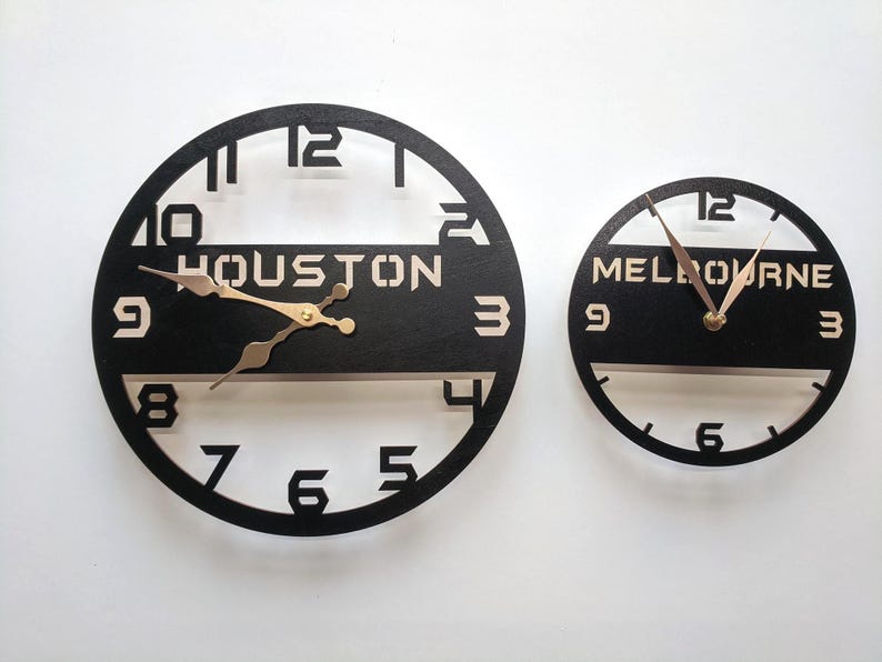 Pu&ograve; includere: Due orologi da parete rotondi neri. Uno indica "HOUSTON" e l'altro "MELBOURNE" in lettere bianche. Le lancette dell'orologio sono dorate. I numeri sono ritagliati dal quadrante nero.