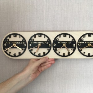 May include: A wooden world time zone clock displaying the time in Los Angeles, Singapore, Madrid, and San Francisco. The clock has a light wood base with four black clock faces and gold hands. The cities' names are printed in white.