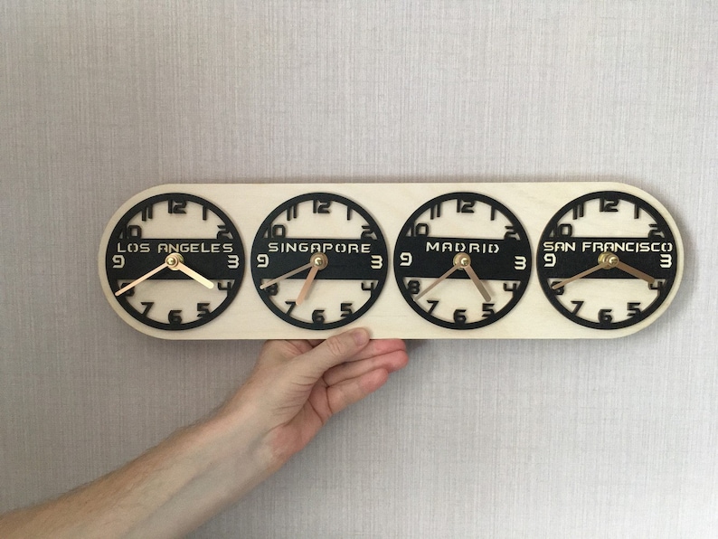 May include: A wooden world time zone clock displaying the time in Los Angeles, Singapore, Madrid, and San Francisco. The clock has a light wood base with four black clock faces and gold hands. The cities' names are printed in white.