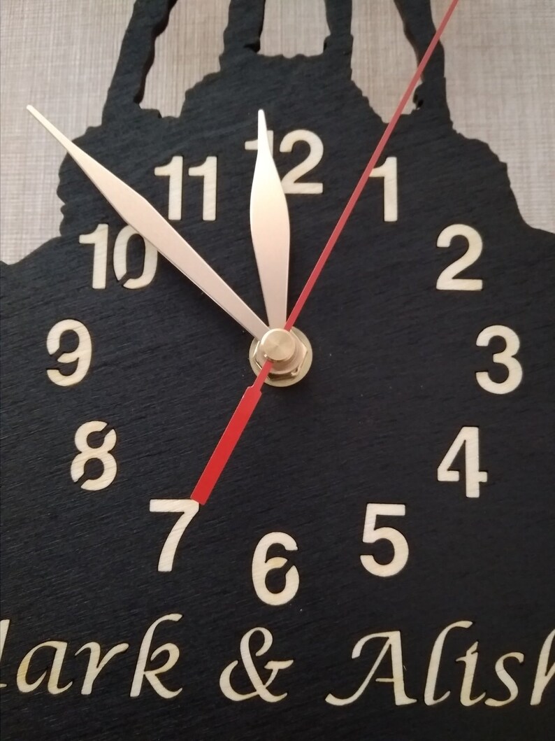 May include: A black wooden clock with white numbers and gold hands. The clock face is shaped like a silhouette of a couple. The clock has the text "Clark & Alish" at the bottom.