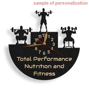 May include: Black wooden clock with a gold clock face and gold hands. The clock face has a silhouette of three people lifting weights. The clock face also has the text "Total Performance Nutrition and Fitness".