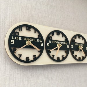 May include: A wooden world time zone clock with four black clock faces displaying the time in Los Angeles, Singapore, Madrid, and San Francisco. The clock hands are light brown, and the clock is mounted on a light-colored wooden base.