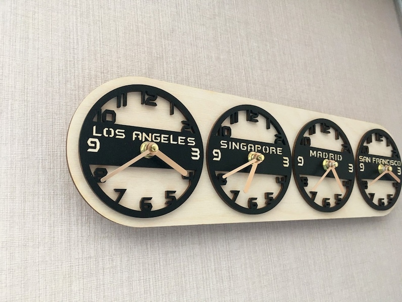 May include: A wooden world time zone clock with four black clock faces displaying the time in Los Angeles, Singapore, Madrid, and San Francisco. The clock hands are light brown, and the clock is mounted on a light-colored wooden base.