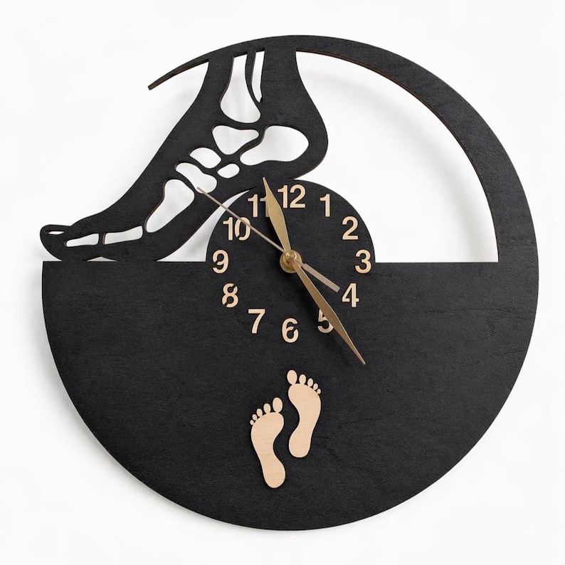 May include: A black wooden wall clock with a unique design. The clock features a cutout of a foot skeleton, a circular clock face with gold hands, and two small wooden footprints. The clock is mounted on a white background.