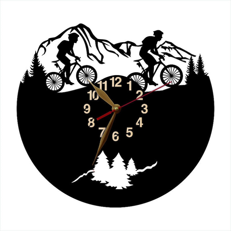 May include: Black wall clock featuring a mountain biking scene. The clock face is black with gold-colored numerals and clock hands. Two cyclists are silhouetted riding bikes in front of a mountain range. The clock has a round shape.