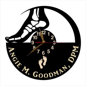 May include: A black and white wall clock featuring a foot skeleton silhouette above a clock face. The clock hands are gold. The clock face has the numbers 1-12. The words "ANGIE M. GOODMAN, DPM" are printed on the clock.