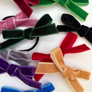 May include: An assortment of velvet hair ties in a range of colours: burgundy, green, black, red, pink, grey, purple, tan, and blue. Each tie is designed with a bow, offering a stylish accessory for any hairstyle.