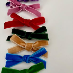 May include: A collection of seven small velvet hair bows arranged in a vertical row. The bows feature a soft, textured finish and are available in a variety of vibrant colours including purple, dusty pink, red, dark green, tan, blue, and bright green.
