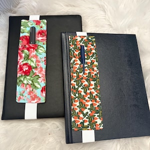 Peut inclure: Deux carnets avec porte-stylos. L'un est noir avec un porte-stylo floral rouge, rose et vert. L'autre est bleu marine avec un porte-stylo à imprimé fraise. Les deux ont un élastique blanc.