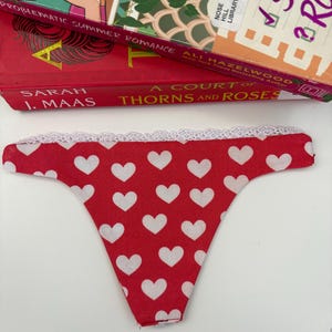 May include: A red thong featuring a repeating pattern of small white hearts. The garment is finished with a delicate white lace trim along the top waistband edge. The fabric is smooth and the design is consistent across the entire front surface.