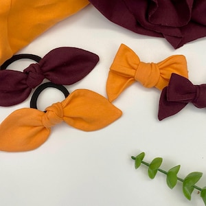 May include: Three fabric hair bows, two orange and one burgundy. The bows are tied with a knot in the centre and have a pointed shape. The bows are on a white surface.