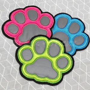 Könnte beinhalten: Drei farbige Pfotenabdruck-Patches. Einer ist rosa, einer blau und einer grün. Jeder Pfotenabdruck hat eine graue Mitte und einen schwarzen Rand. Die Patches befinden sich auf einem grau-weißen geometrischen Hintergrund.