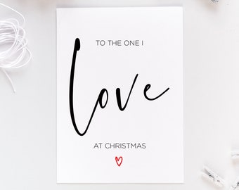 May include: A white Christmas card with the message "TO THE ONE I love AT CHRISTMAS" in black script, with a red heart. The card is accompanied by a wooden bead garland and a silver star decoration, all on a white surface.
