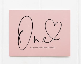 Puede incluir: Tarjeta rosa con caligrafía negra que dice "one" con un corazón dibujado en el bucle de la "e". El texto de abajo dice "Happy First Birthday Ariel!"