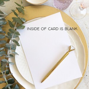 May include: A blank white card with the text "INSIDE OF CARD IS BLANK" sits on a white plate, next to a gold pen with a diamond-shaped top. A gold ring and a glass of pink liquid are also visible.