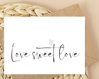 May include: A white card with the words "Love sweet Love" in black script, topped with a small heart. The card is inside a brown envelope, set on a woven mat with a beige background.
