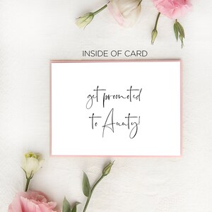 May include: A white card with pink trim and the text "get promoted to Aunty!" written in black cursive. The card is surrounded by pink and white flowers.