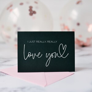 Może przedstawiać: Czarna kartka z ręcznie napisanym białym tekstem "I just really, really love you" z małym sercem narysowanym obok słowa "you". Kartka leży na r&oacute;żowej kopercie na białym marmurowym tle. W tle widoczne są przezroczyste balony z r&oacute;żowymi i złotymi konfetti.