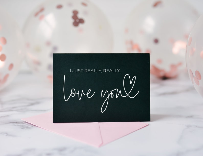 Może przedstawiać: Czarna kartka z ręcznie napisanym białym tekstem "I just really, really love you" z małym sercem narysowanym obok słowa "you". Kartka leży na r&oacute;żowej kopercie na białym marmurowym tle. W tle widoczne są przezroczyste balony z r&oacute;żowymi i złotymi konfetti.