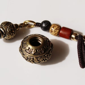 May include: A brass bottle stopper with intricate designs and a beaded tassel. The stopper is made of two pieces, a round top with a hole in the center and a smaller, cylindrical bottom piece. The tassel is made of brown cord and wooden beads.