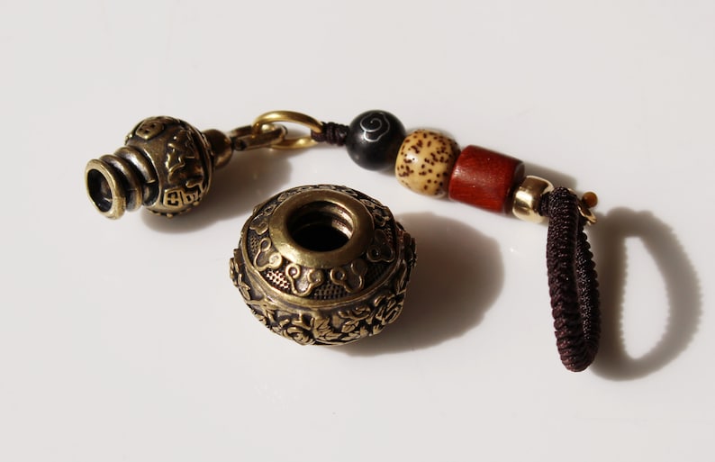 May include: A brass bottle stopper with intricate designs and a beaded tassel. The stopper is made of two pieces, a round top with a hole in the center and a smaller, cylindrical bottom piece. The tassel is made of brown cord and wooden beads.
