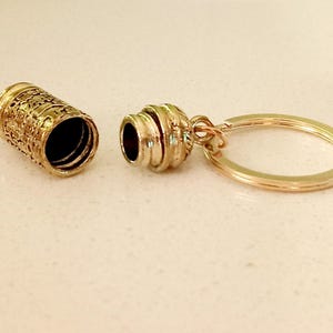 May include: Gold-toned metal keychain with a small, cylindrical container that opens and closes. The container has a decorative, textured design.