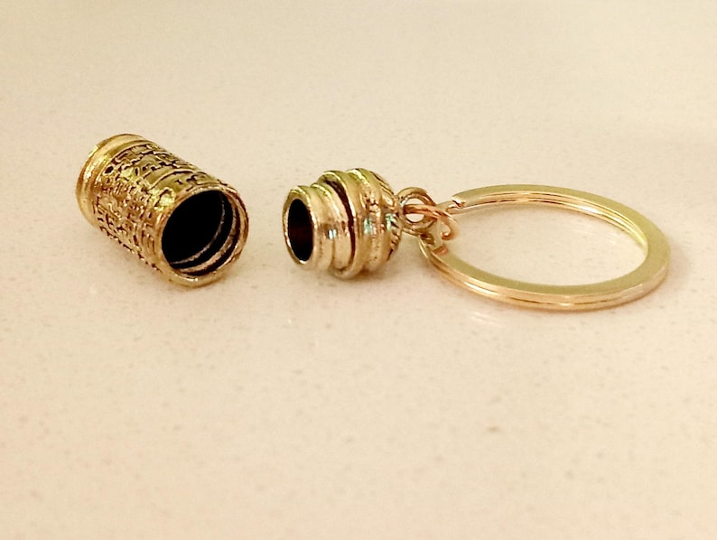 May include: Gold-toned metal keychain with a small, cylindrical container that opens and closes. The container has a decorative, textured design.