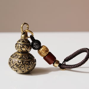 May include: A brass gourd-shaped keychain with intricate floral designs and Chinese characters. The keychain has a brown cord with wooden and black beads.