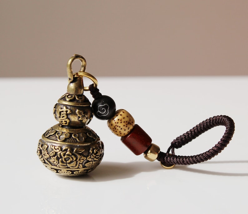 May include: A brass gourd-shaped keychain with intricate floral designs and Chinese characters. The keychain has a brown cord with wooden and black beads.