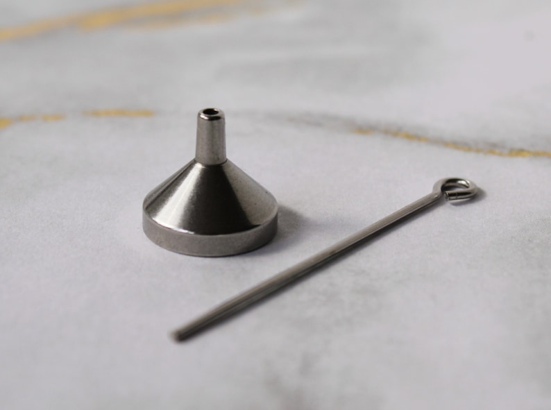 May include: A small, silver funnel with a long, thin, silver rod with a loop at one end. The funnel and rod are on a white surface.