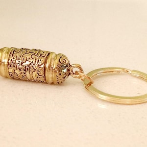 May include: Gold-colored metal keychain with a small, cylindrical charm that has intricate engravings. The charm is attached to a gold-colored metal ring.