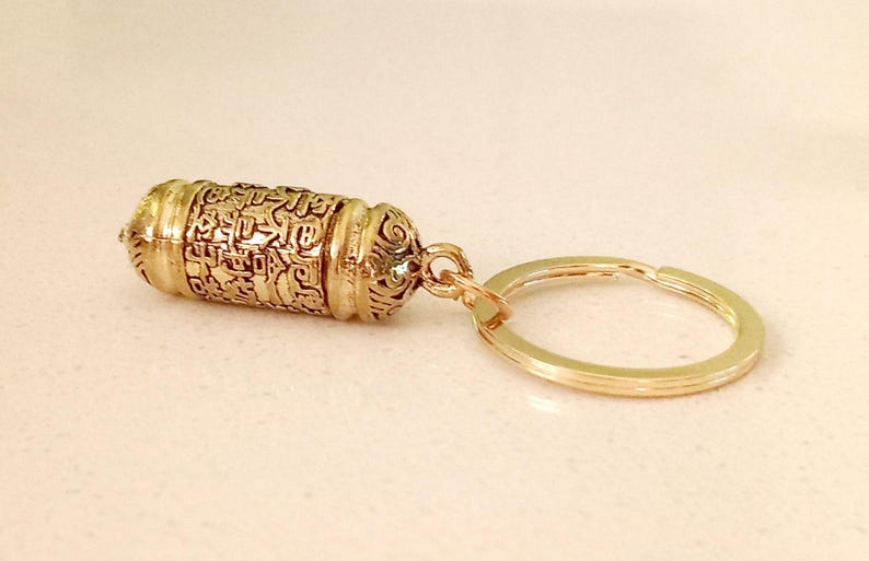 May include: Gold-colored metal keychain with a small, cylindrical charm that has intricate engravings. The charm is attached to a gold-colored metal ring.