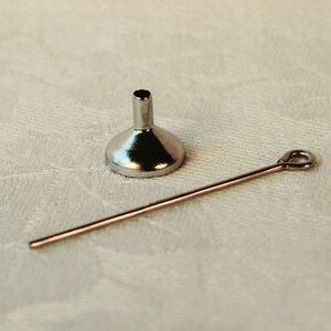 May include: A silver funnel with a long, thin, silver rod attached to it. The funnel and rod are lying on a white surface.
