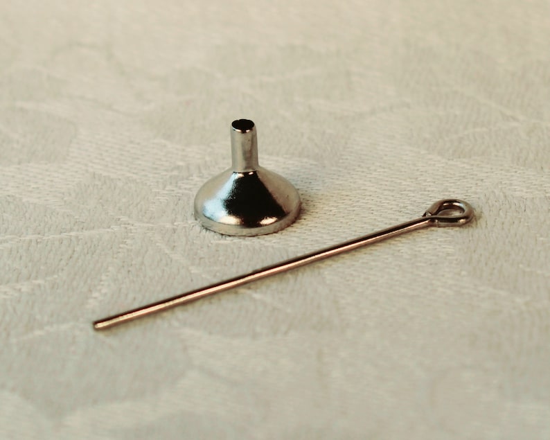 May include: A silver funnel with a long, thin, silver rod attached to it. The funnel and rod are lying on a white surface.