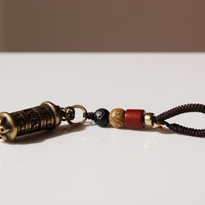 May include: A small, brass prayer wheel with intricate carvings and a brown cord with wooden and black beads. The prayer wheel is attached to the cord with a gold-colored ring.