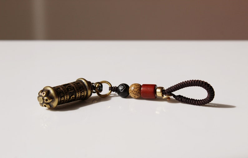 May include: A small, brass prayer wheel with intricate carvings and a brown cord with wooden and black beads. The prayer wheel is attached to the cord with a gold-colored ring.