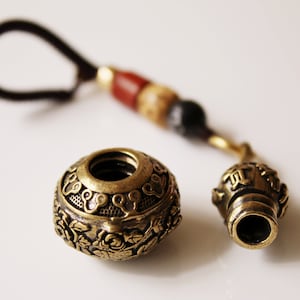 May include: A brass key chain with a round, ornate charm and a smaller, cylindrical charm. The charms are attached to a black cord with red, brown, and black beads.