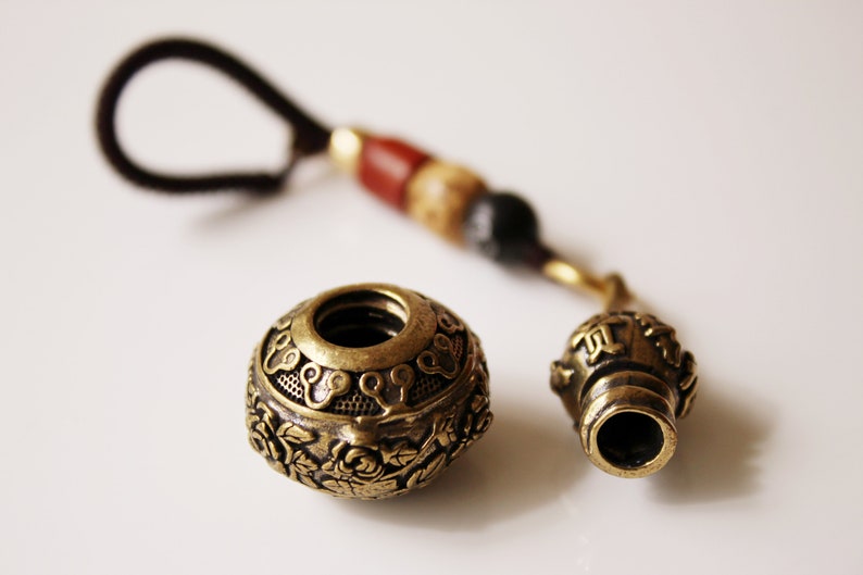May include: A brass key chain with a round, ornate charm and a smaller, cylindrical charm. The charms are attached to a black cord with red, brown, and black beads.